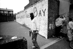 A hooded paramilitary gunman stands in front of a painted wall that reads "You Are Now In Loyalist Tiger Bay." BELFAST, NORTHERN IRELAND, 1990. Ⓒ Ed Kashi