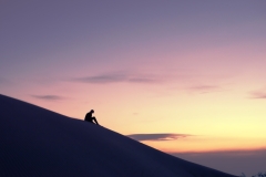 Lone woman contemplating the horizon in the desert from atop a dune against a golden sky at sunset.
