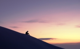 Lone woman contemplating the horizon in the desert from atop a dune against a golden sky at sunset.
